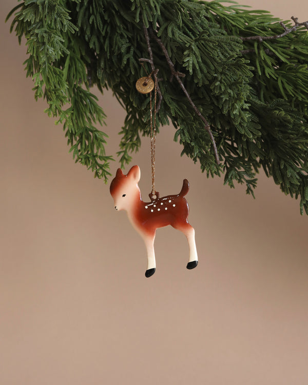 The Maileg Metal Bambi Deer Ornament, featuring white spots and a gold chain, hangs on a green pine branch against a beige background—ideal for Christmas tree décor or adding charm to your home.