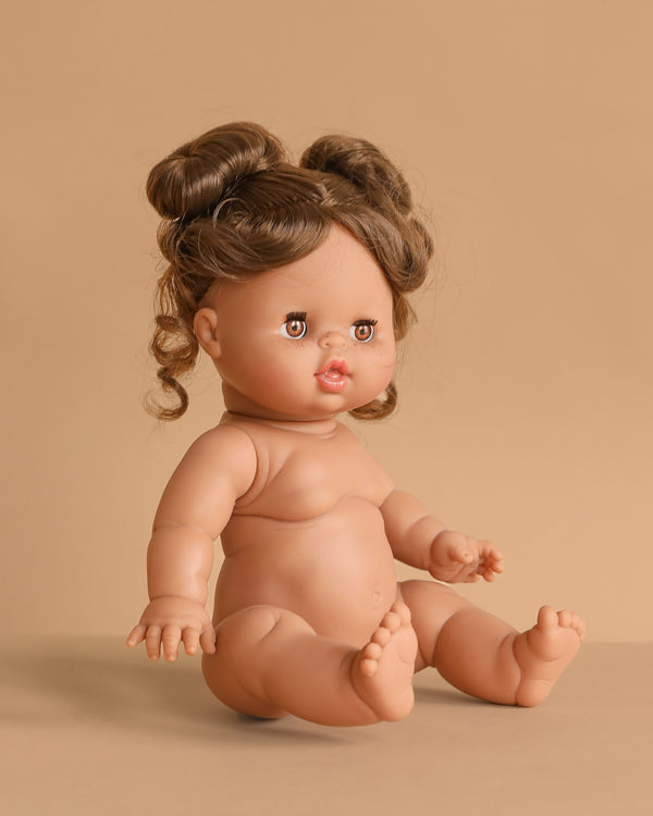 The 13" Minikane Baby Doll With Sleeping Eyes - Madeleine sits on a beige surface with brown hair in two buns. The anatomically correct doll features chubby limbs and a slightly open mouth. Clothing sold separately.