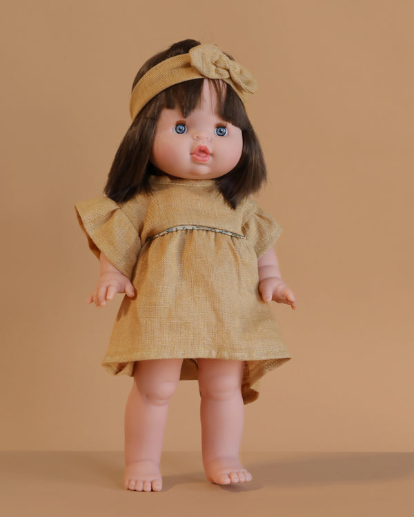 The Minikane Standing Doll (14") - Chléa With Clothing features straight dark brown hair, blue eyes, a tan dress with matching headband, and stands barefoot against a solid light brown background.
