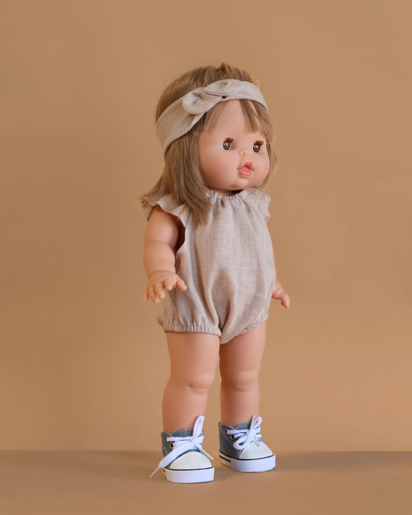 The Minikane Standing Doll (14") - Zoelie With Clothing & Shoes features light brown hair with a matching headband, beige romper, and gray sneakers. The anatomically correct doll stands with arms slightly raised against a tan background. Made in Spain.