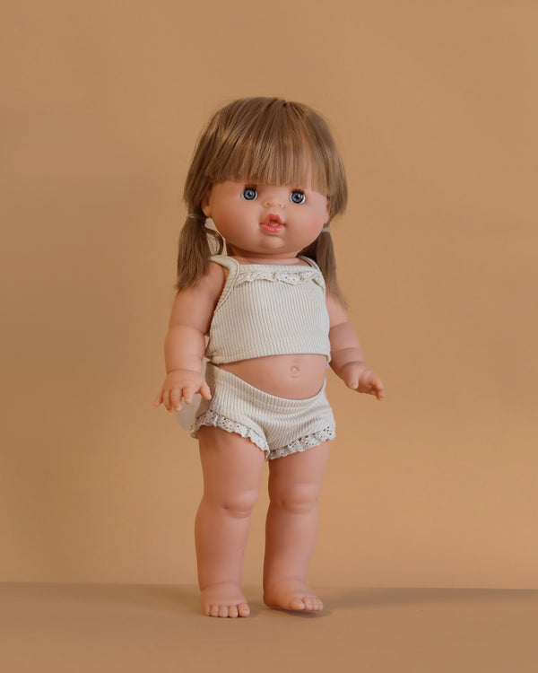The Minikane Standing Doll (14") - Yzé With Clothing features straight light brown hair, blue eyes, and bangs. This anatomically correct doll stands upright wearing a cream tank top and matching shorts against a plain light brown background.