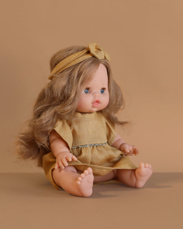 The Minikane Baby Doll (13") - Eleanor With Clothing features long light brown hair, blue eyes, and sits barefoot in a yellow dress with matching headband against a beige background, legs outstretched.