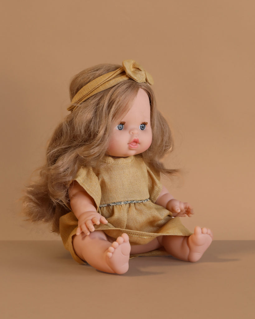 The Minikane Baby Doll (13") - Eleanor With Clothing features long light brown hair, blue eyes, and sits barefoot in a yellow dress with matching headband against a beige background, legs outstretched.
