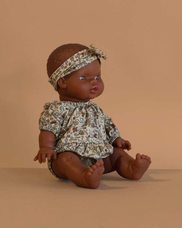 The Minikane Doll (13") - Zélie With Clothing, a dark-skinned, anatomically correct doll dressed in a floral outfit and matching headband, sits on a beige surface with a plain beige background. Made in Spain.