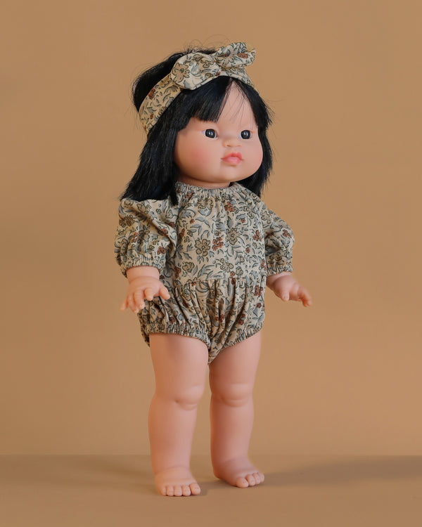 The Minikane Standing Doll (14") - Jade With Clothing features long black hair, a floral romper with matching headband, and stands upright against a beige background. This anatomically correct doll is beautifully made in Spain.