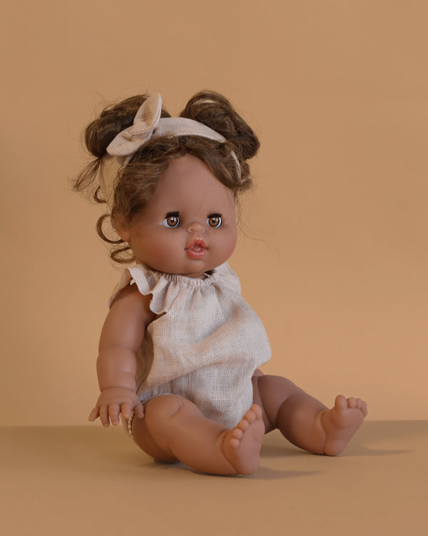 The Minikane Doll With Sleeping Eyes (13") - Madeleine With Clothing, featuring brown hair in a top knot and a beige sleeveless outfit with headband, sits against a beige background. This anatomically correct doll is perfect for imaginative play.