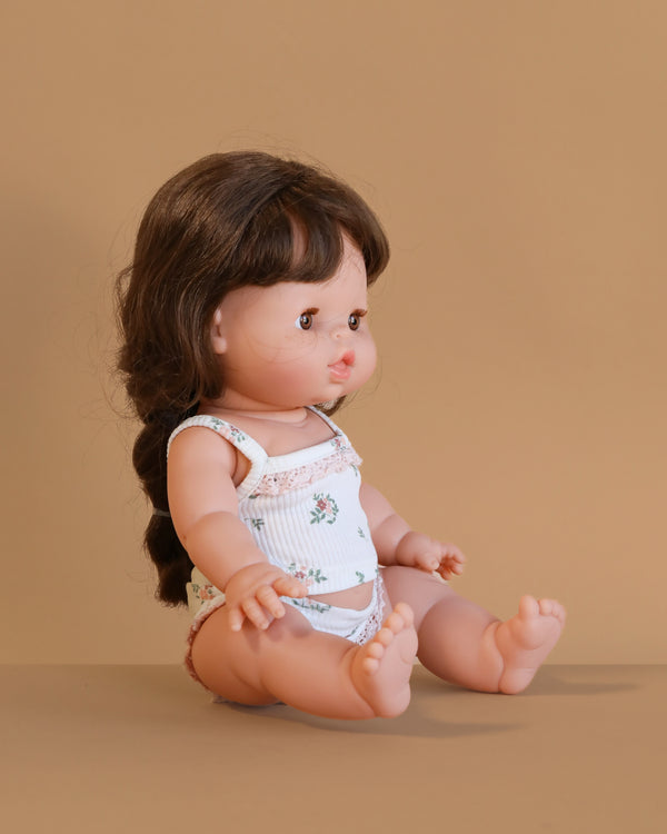 The Minikane Doll (13") - Valentine With Clothing, featuring long brown braided hair and a white floral outfit, sits on a beige surface against a beige background, facing left.