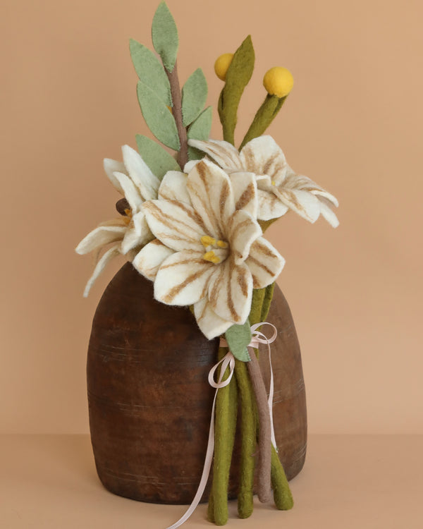 The Handcrafted Felt Flower Bouquet features white and yellow wool felt blooms with green stems arranged in a brown vase, accented by pale ribbons, and set against a plain beige background.
