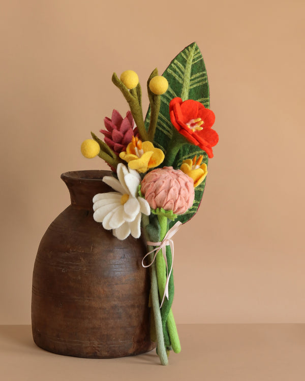 A Handcrafted Felt Flower Bouquet of red, white, pink, and yellow blooms tied with string sits beside a rustic brown clay vase, showcasing sustainable flowers against a beige background.