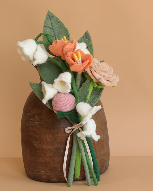 The Handcrafted Felt Flower Bouquet features peach, pink, and white felt flowers with green leaves in a brown vase tied with a pale ribbon, perfect for sustainable home decor. Displayed against a beige background.
