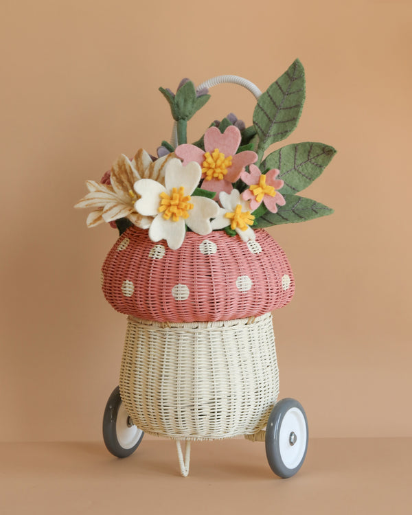 The Basket of Felt Flowers is a small wicker basket on wheels with a handle, topped by a pink polka-dot cushion and filled with eco-friendly felt flowers and large green leaves, shown on a tan background.