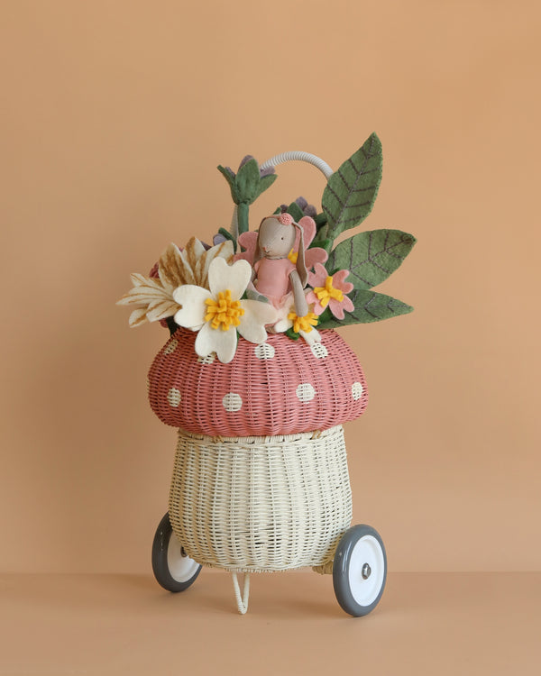 A whimsical wicker Basket of Felt Flowers with Maileg Bunny on wheels, shaped like a mushroom and adorned with felt flowers and large green leaves, set against a plain beige background.