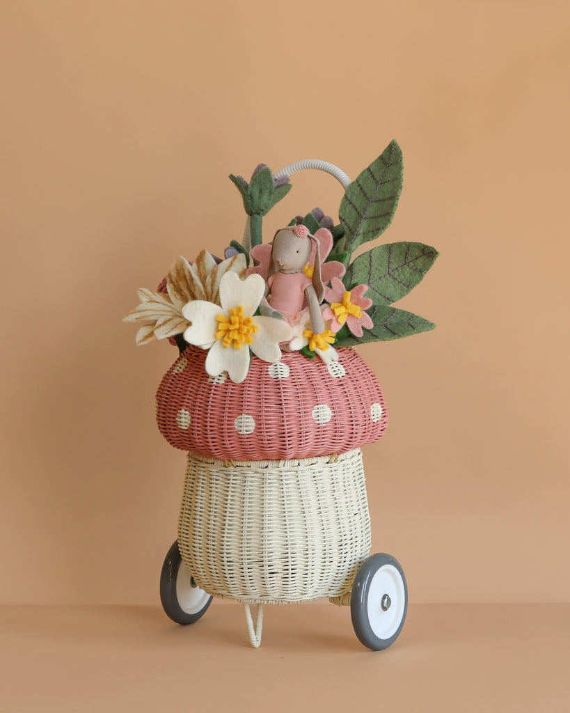 A whimsical wicker Basket of Felt Flowers with Maileg Bunny on wheels, shaped like a mushroom and adorned with felt flowers and large green leaves, set against a plain beige background.
