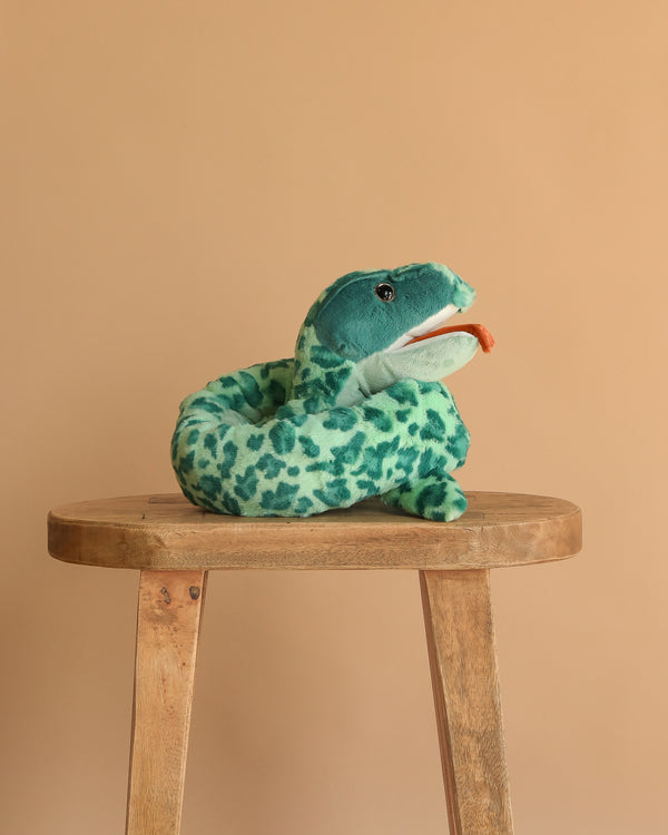 The Steiff Gin Snake Stuffed Plush Animal, 51 Inches, featuring soft green spotted plush and a red tongue, sits coiled on a wooden stool against a plain beige background.
