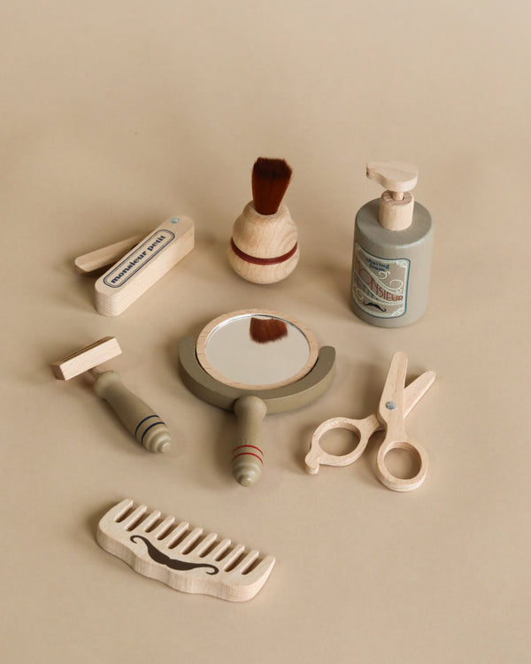 A collection of non-toxic toys including a brush, razor, toothbrush, mirror, scissors, comb, and a dispenser from the Pretend Play Wooden Barber Set arranged on a beige surface.