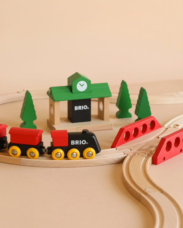 The BRIO World Train Set Classic Figure 8 Set features classic red and black trains on curved tracks, with red bridges, green pine trees, and a green station—crafted in timeless Scandinavian design.