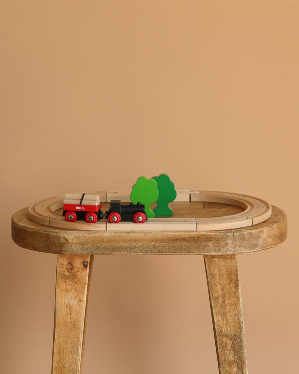 The BRIO Little Forest Train Set features a wooden toy train with red wheels on a wooden track, passing two green trees on a wooden table, all set against a plain beige background.