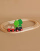 The BRIO Little Forest Train Set features a wooden toy train with red wheels on an oval track, hauling small cargo. Two green trees decorate the center, creating a classic scene against a light brown background.