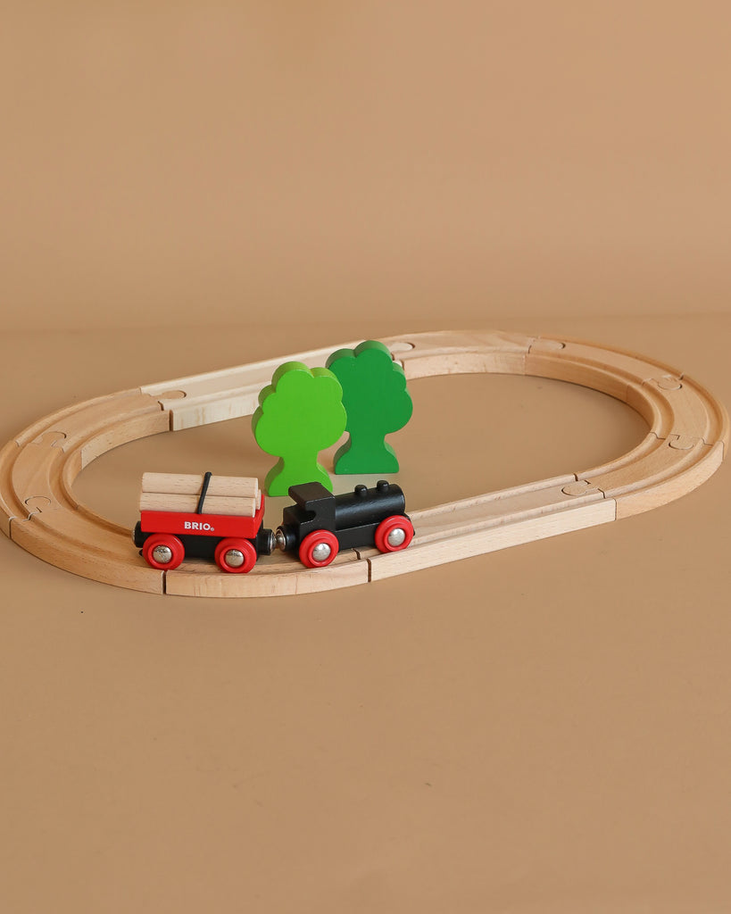 The BRIO Little Forest Train Set features a wooden toy train with red wheels on an oval track, hauling small cargo. Two green trees decorate the center, creating a classic scene against a light brown background.