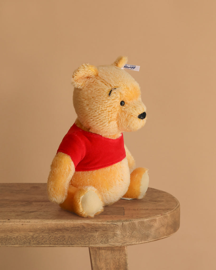 The Steiff Disney’s Winnie the Pooh Open Edition Collectible is a 10-inch plush bear with yellow fur and a red shirt, shown sitting upright on a wooden bench against a plain beige background.