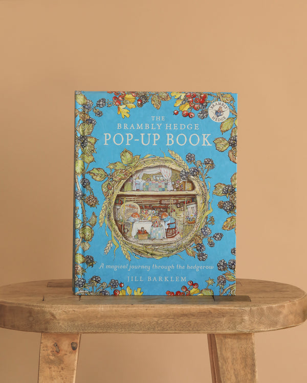 The Brambly Hedge Pop-Up Book by Jill Barklem stands upright on a wooden table against a beige background, its cover featuring vibrant illustrations of a mouse home and foliage on a blue backdrop.