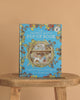 The Brambly Hedge Pop-Up Book by Jill Barklem stands upright on a wooden table against a beige background, its cover featuring vibrant illustrations of a mouse home and foliage on a blue backdrop.