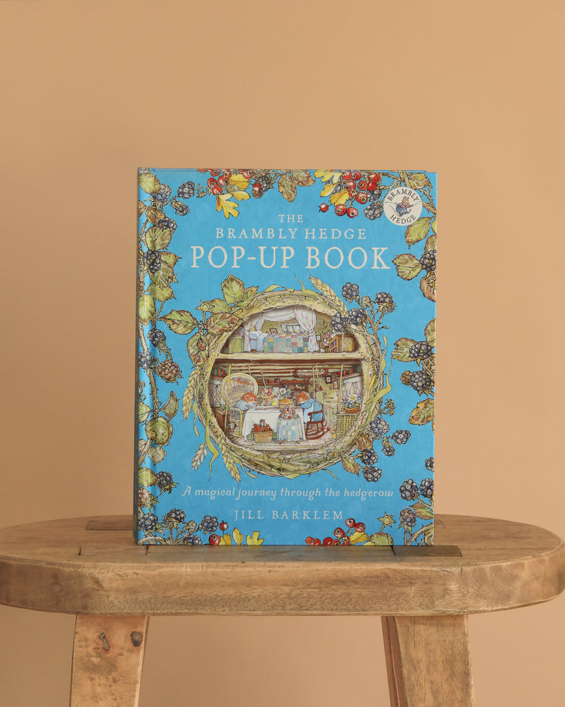 The Brambly Hedge Pop-Up Book by Jill Barklem stands upright on a wooden table against a beige background, its cover featuring vibrant illustrations of a mouse home and foliage on a blue backdrop.