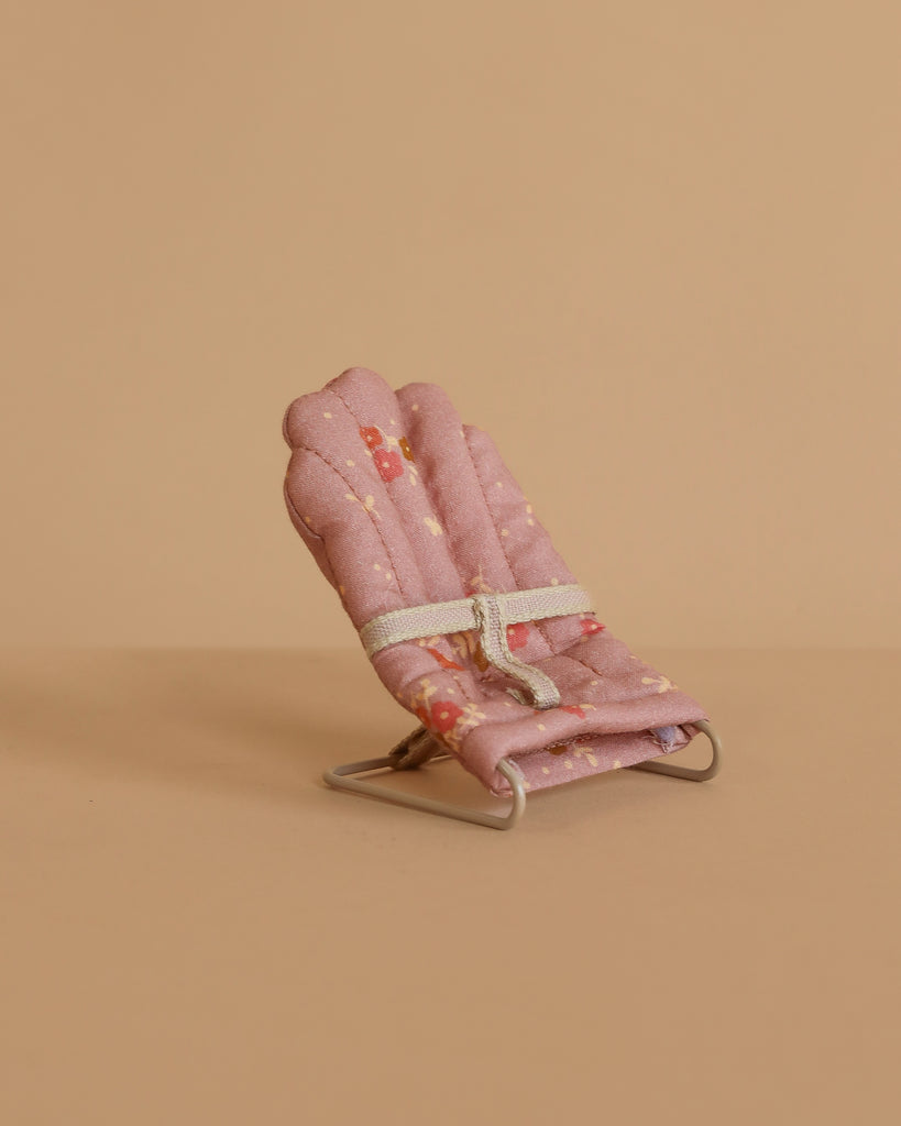The Maileg Baby Twins with Babysitters Set features a small, pink padded chair with a white strap—perfect for Maileg baby mice—set against a beige background. Its subtle patterned seat and sturdy metal frame suit any dollhouse decor.