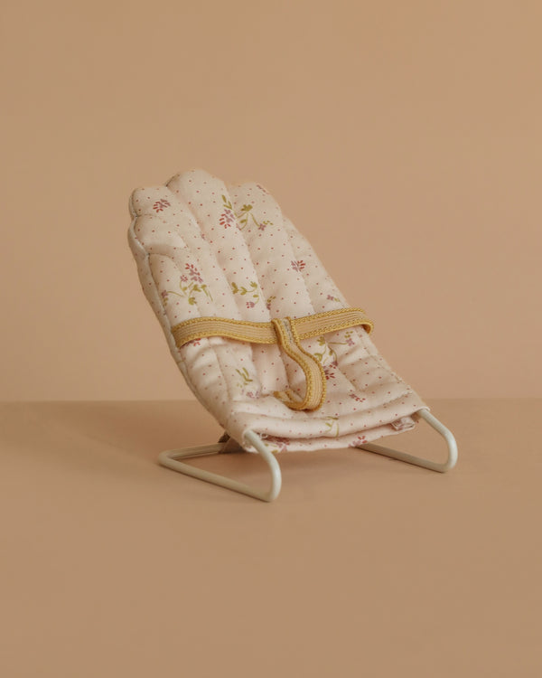 The Maileg Babysitter, Micro - Off White features a small, padded seat with a floral pattern and yellow safety straps on a white metal frame. Perfect as baby doll furniture or for micro-sized rabbits; displayed on a beige surface and background.