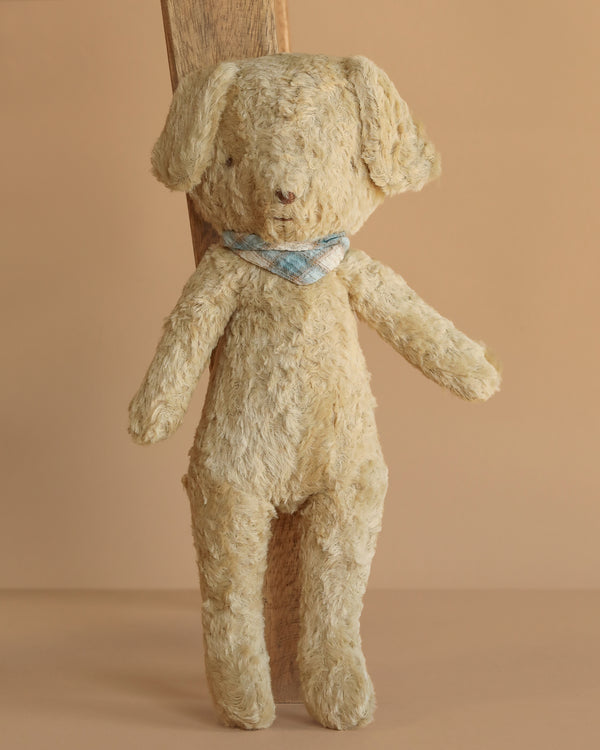 The Maileg Poodle Dog, Vintage Plush - Soft Sand stands upright with floppy ears and a blue and white checkered scarf. Made from cotton viscose plush, it’s displayed against a simple wooden plank and tan background.