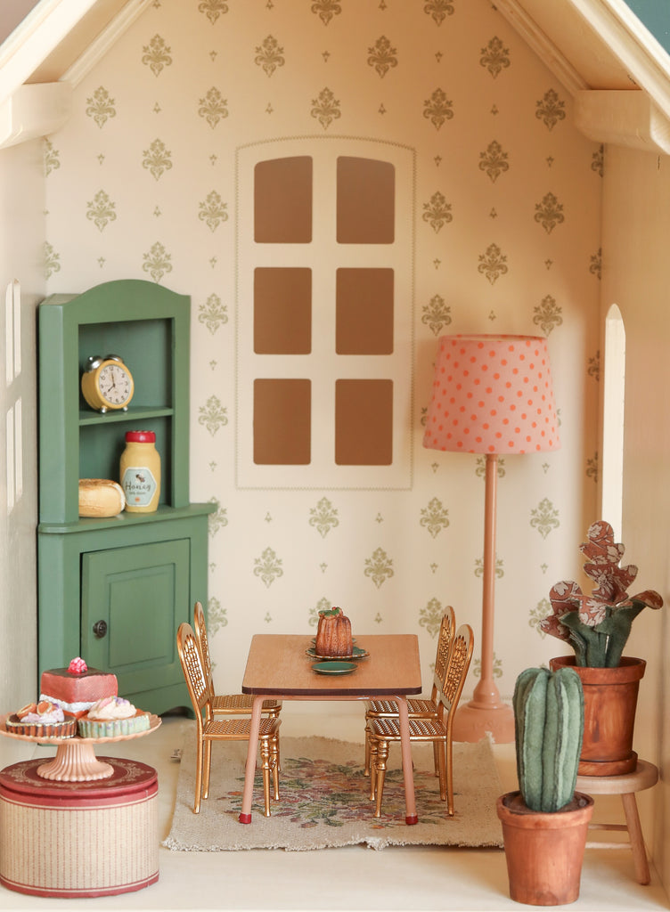 The Maileg Fully Furnished Dollhouse Set + Surprise Gift includes a charming dining room scene with a table, four chairs, green cabinet with dishes, pink polka-dot lamp, cakes on display, potted plants, and patterned wallpaper.