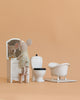 A plush mouse in pajamas stands on a stool, gazing into a vanity mirror beside the Maileg Little Bother Bathroom Set and Maileg furniture, all set against a peach backdrop inspired by the Mouse Hole Farmhouse.