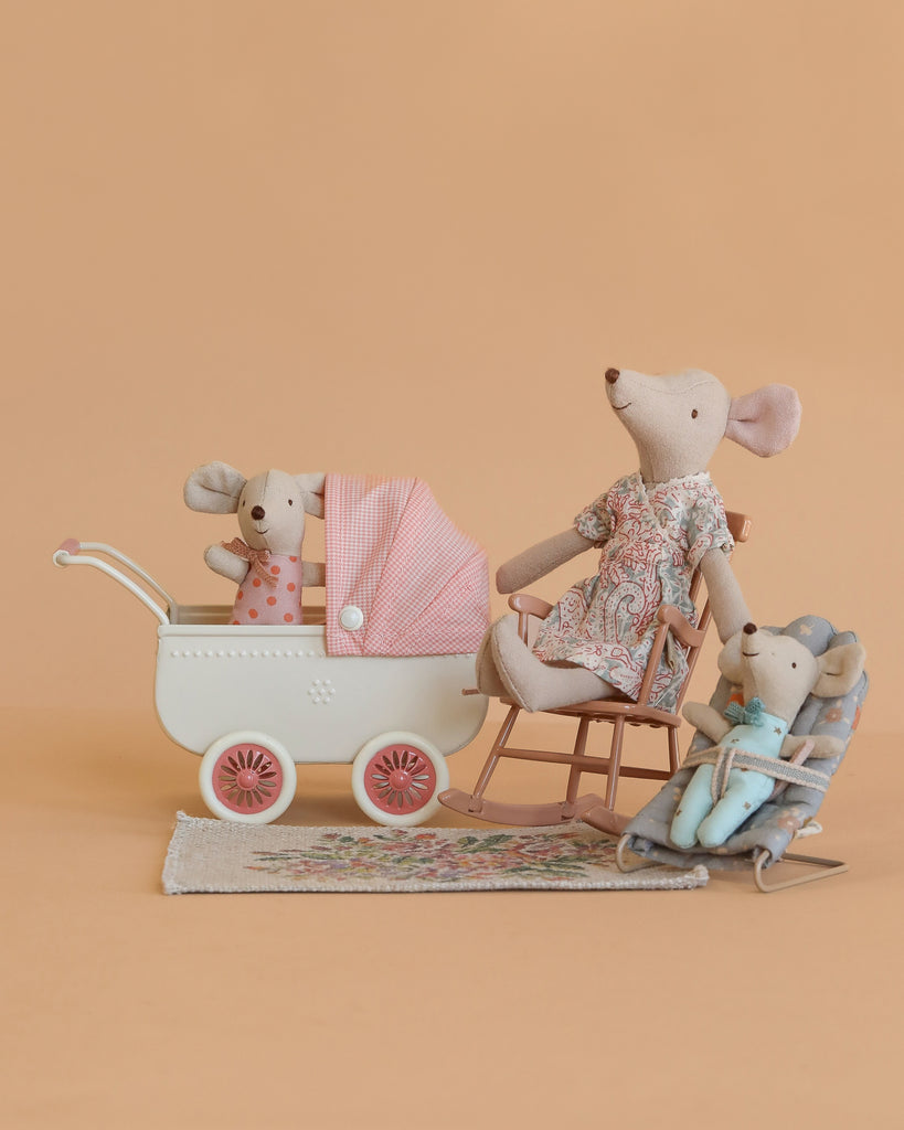 The Maileg Nursery Set features three fabric toy mice arranged as a family with nursery furniture—a rocking chair, bouncer, and baby carriage with pink blanket—displayed on a patterned rug against a peach background.