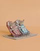 The Maileg Baby Twins with Babysitters Set features two baby mice in bathrobes relaxing on pink and blue chairs atop a dollhouse rug, all set against a plain peach background.