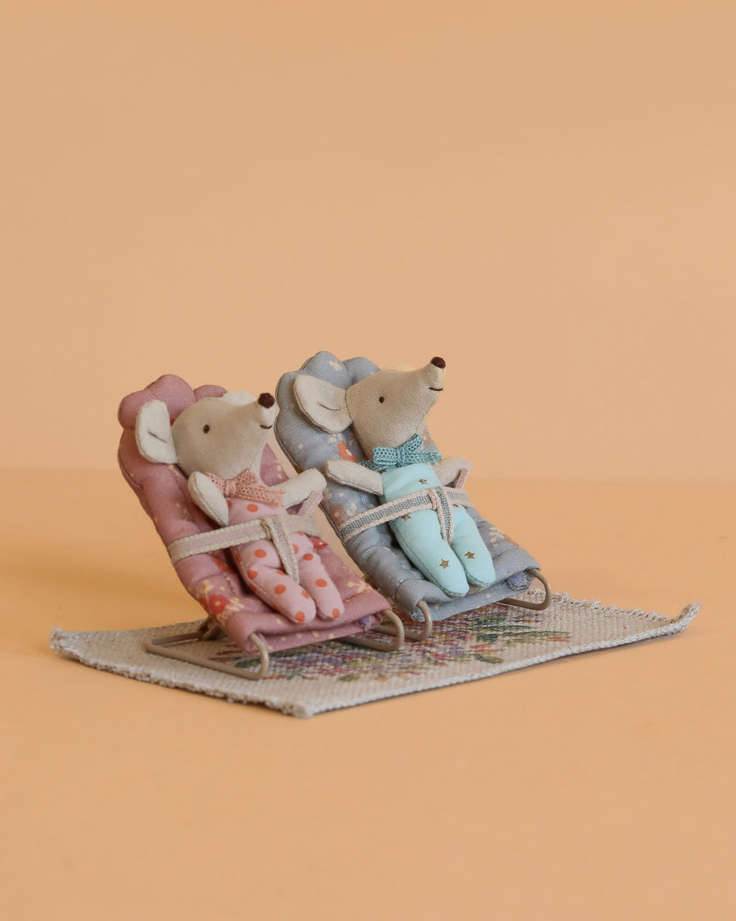 The Maileg Baby Twins with Babysitters Set features two baby mice in bathrobes relaxing on pink and blue chairs atop a dollhouse rug, all set against a plain peach background.
