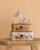 The Maileg Unicorn with Suitcase Set features a plush unicorn sitting on two floral and animal-print suitcases, all displayed on a wooden table with a peach-colored background.