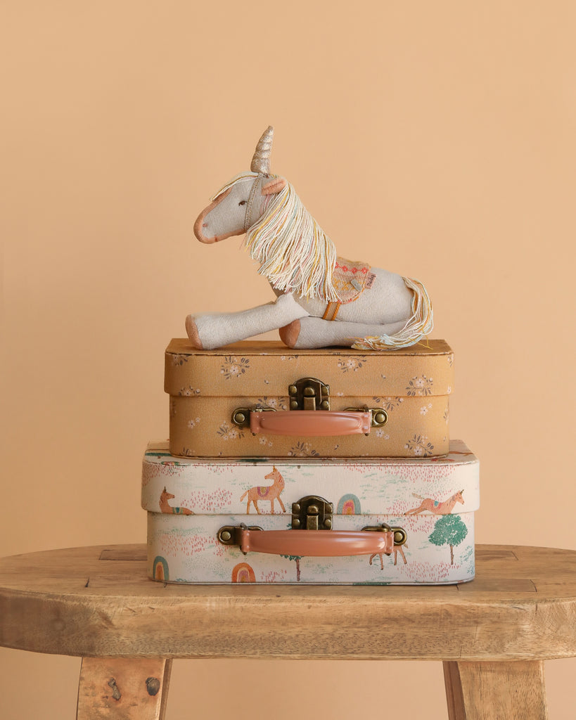 The Maileg Unicorn with Suitcase Set features a plush unicorn sitting on two floral and animal-print suitcases, all displayed on a wooden table with a peach-colored background.