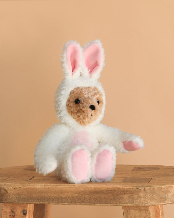 The Steiff Teddy Bear with Bunny Hoodie Costume, 11 inches, features a brown teddy dressed in a fluffy white bunny hoodie with pink ear and paw details, sitting on a wooden stool against a beige background.