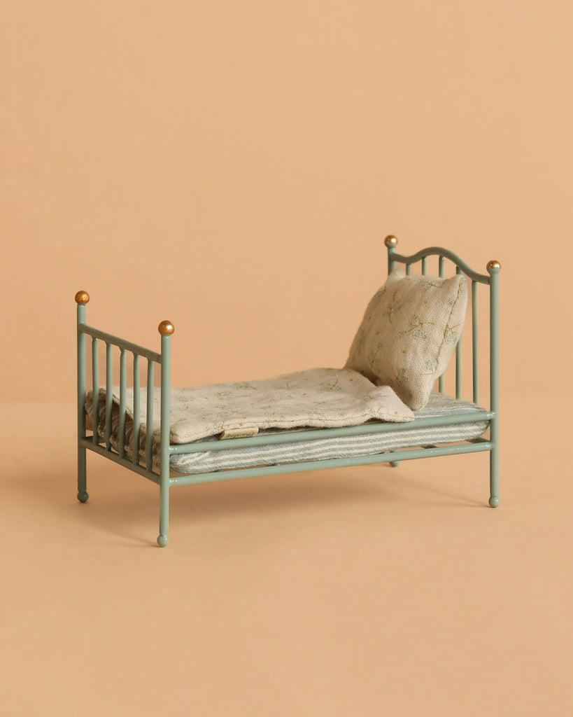 The Maileg Little Sister Bedroom Set features a small mint green metal toy bed with pillow and blanket beside a Maileg Vintage Floor Lamp, all placed against a plain peach-colored background.