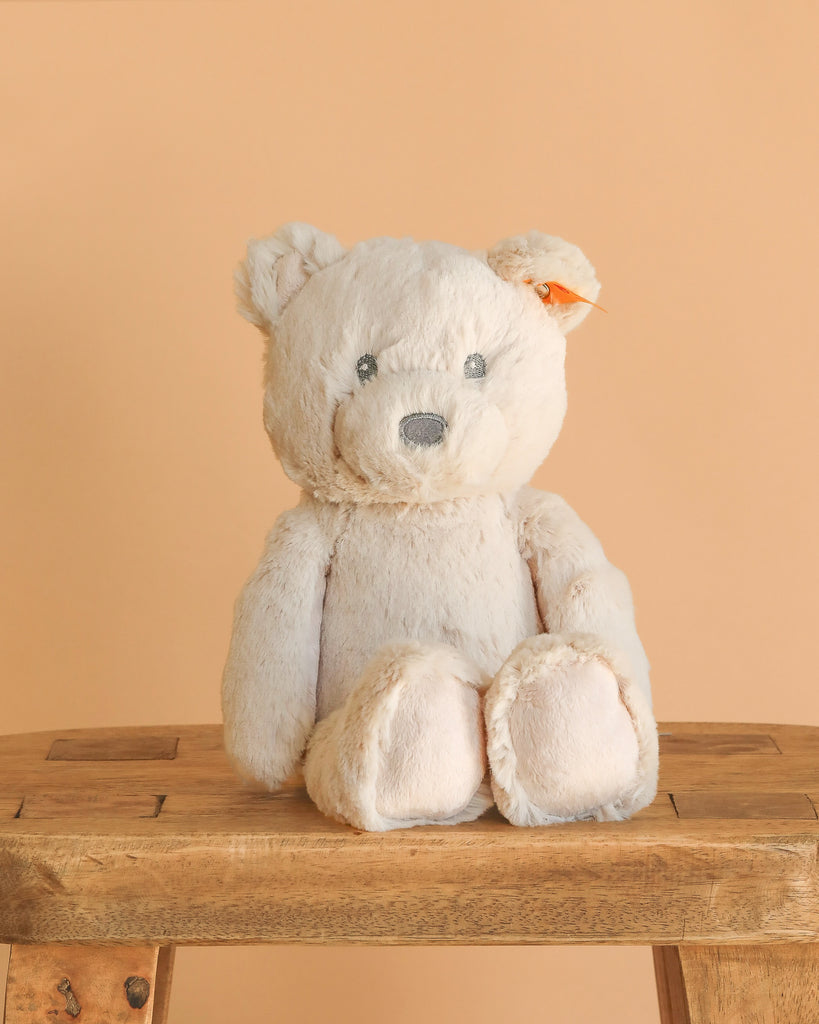 The Steiff Bearzy Teddy Bear Stuffed Plush for Baby, 11 inches, in light beige with gray eyes and nose, made in Germany from ultra soft plush, sits upright on a wooden stool against a peach background.