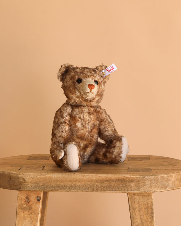 The Steiff 1926 Mohair Teddy Bear, made in Germany, with a red ear tag, sits on a wooden stool against a plain beige background.