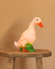 The Duck Lamp, with its orange beak and green grass base, adds a whimsical touch to any child's room—displayed here on a rustic wooden stool against a plain beige background.