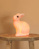 The White bunny Lamp, with its smooth white surface and adorable features, sits on a rustic wooden stool against a beige background, casting a soft, warm glow as a whimsical night light for any nursery.