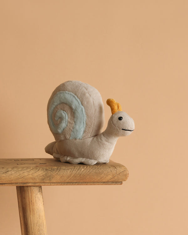 The Steiff Snaily Snail Stuffed Plush Animal, 8 inches, with a light gray body, yellow antennae, and blue spiral shell, sits on a wooden stool against a plain beige background.