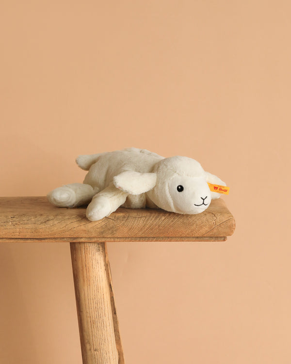 The Steiff Floppy Linda Lamb plush stuffed baby toy, 8 inches, with a yellow tag, rests on a rustic wooden bench against a plain beige background.