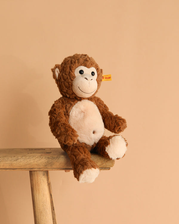 The Bodo Monkey Plush Stuffed Toy, 12 Inches, in brown and white with a stitched smile, sits on a wooden bench against a beige background. A yellow tag in its ear marks it as an authentic Steiff product.