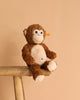 The Bodo Monkey Plush Stuffed Toy, 12 Inches, in brown and white with a stitched smile, sits on a wooden bench against a beige background. A yellow tag in its ear marks it as an authentic Steiff product.