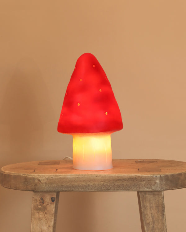 The Small Red Mushroom Lamp, featuring a glowing red cap and white stem, adds a whimsical touch as it sits on a rustic wooden table against a plain beige wall.