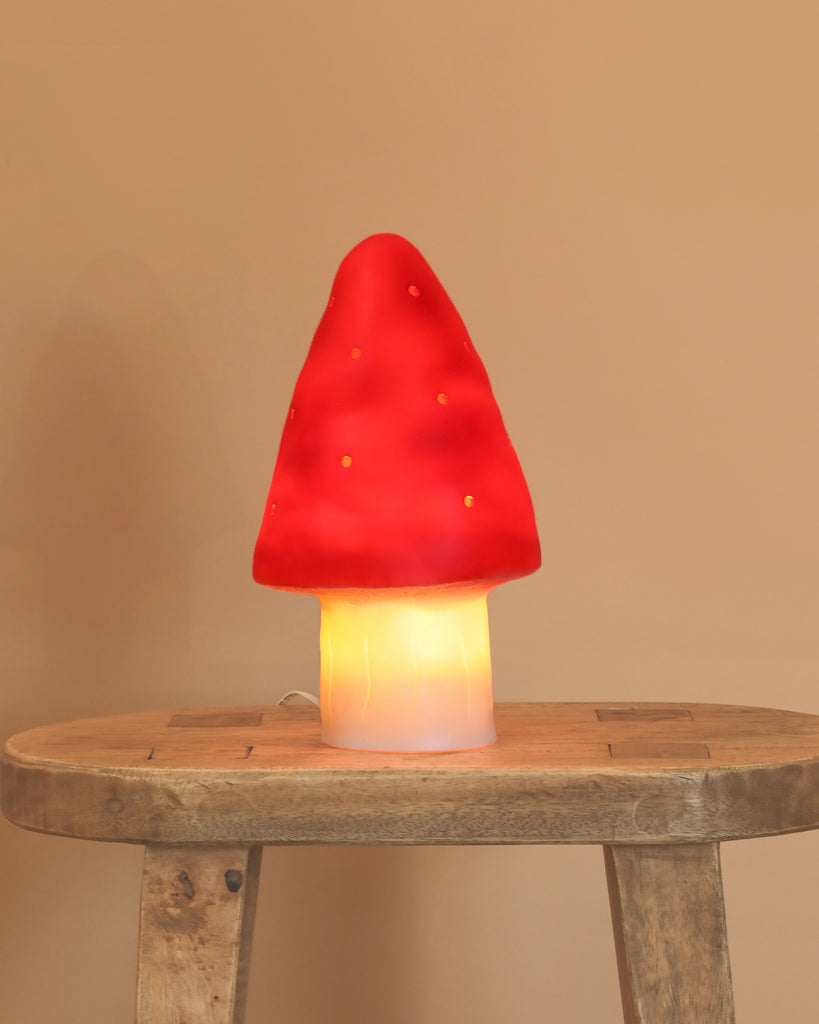 The Small Red Mushroom Lamp, featuring a glowing red cap and white stem, adds a whimsical touch as it sits on a rustic wooden table against a plain beige wall.