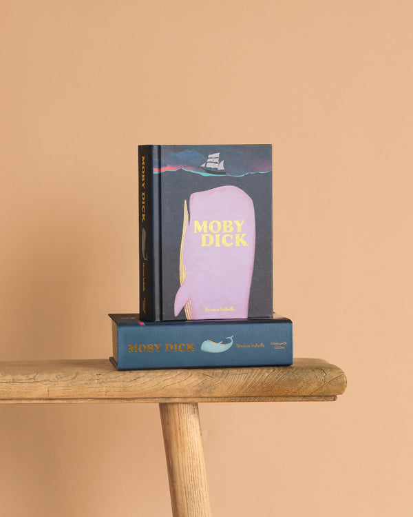 A "Moby Dick | Melville | Collector's Edition | Hardcover" stands upright on a wooden bench, with another copy of the same book lying flat beneath it against a plain light beige background.